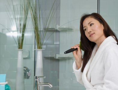 A woman is brushing her teeth in front of a mirror, showcasing her Loxxie Hair while maintaining her oral care.