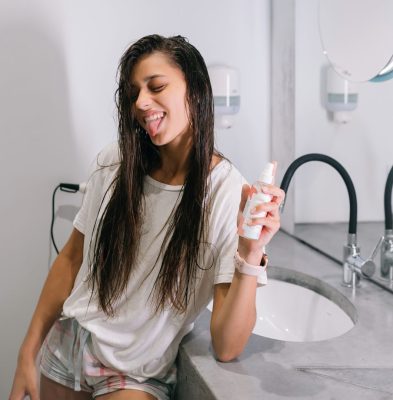 A woman is brushing her teeth in front of a mirror, showcasing her Loxxie Hair while maintaining her oral care.